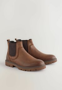 Braune Chelsea-Stiefel aus Leder mit elastischen Seitenpaneelen und dicken Gummisohlen, die nebeneinander auf einem hellen Hintergrund positioniert sind.