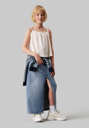Weißes geripptes Cropped-Top mit dünnen Trägern, blauer Karosweater um die Taille gebunden und einem langen blauen Jeansrock mit einem seitlichen Schlitz. Weiße Sneakers.