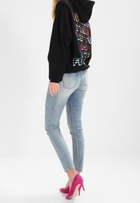 Sudadera con capucha negra y texto multicolor en la parte posterior, combinada con jeans ajustados de color azul claro y zapatos de tacón alto rosas.