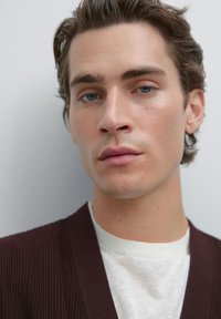 Giovane uomo con occhi azzurri e capelli castani stilizzati, indossa una camicia bianca sotto un cardigan scuro a coste, su uno sfondo grigio chiaro uniforme.