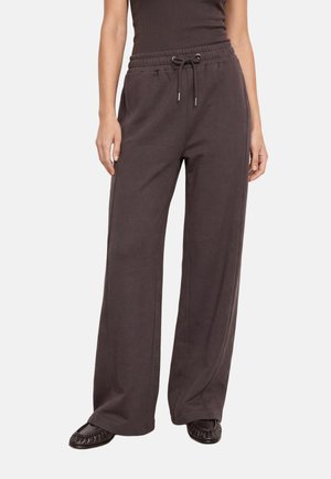 Femme portant un pantalon de jogging marron foncé à jambes larges avec taille à cordon et des chaussures plates marron foncé, debout, les mains le long du corps.