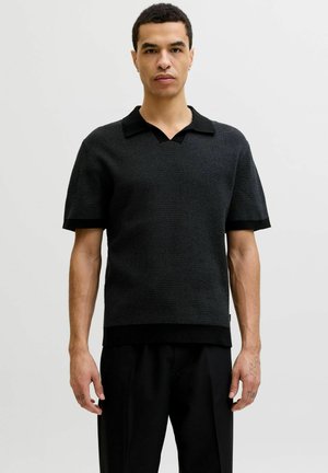 Młody mężczyzna stoi, ubrany w czarną, krótkorękawową, teksturowaną, dzianą koszulkę polo i czarne spodnie na jednolitym jasnym tle.