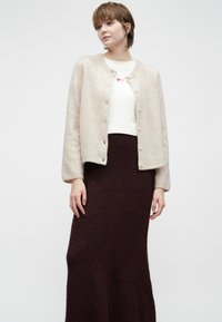 Vrouw in een beige cardigan over een wit shirt met twee kleine rode harten, gecombineerd met een donker bordeauxlange rok, staand tegen een witte achtergrond.