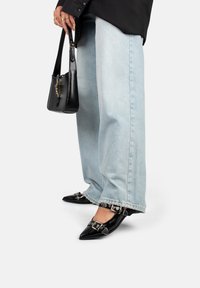 Zapatos de piel negra de punta con detalles de hebilla, combinados con jeans de pierna ancha de color azul claro y un pequeño bolso negro.