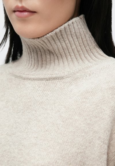 Γυναίκα που φοράει μπεζ ριγωτό πουλόβερ με ριχτό λαιμό, με σκούρα, ίσια μαλλιά που φαίνονται μερικώς.