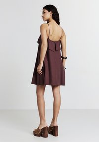 Bordeauxfarbenes Kleid mit dünnen Trägern und einem Rüschendetail auf der Rückseite, aus leichtem Stoff gefertigt. Kombiniert mit braunen, Absatz-Mules mit Nieten.