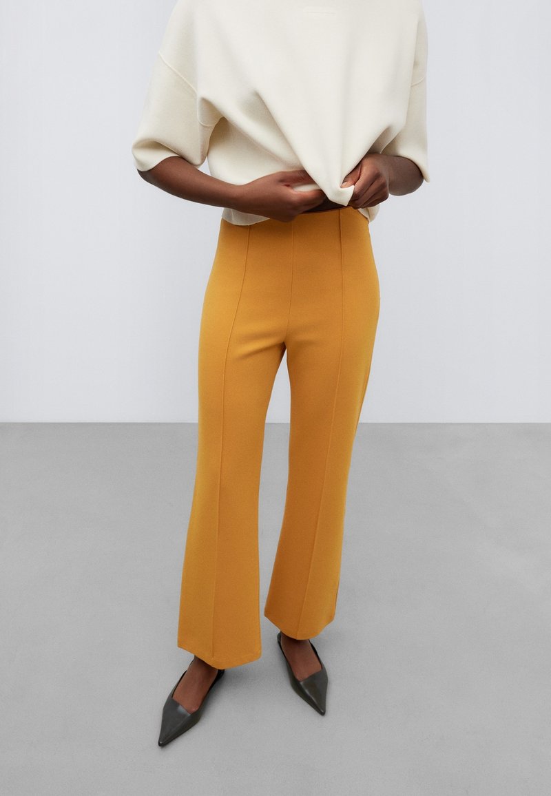 Personne vêtue d’un pantalon évasé taille haute jaune moutarde, d’un haut ample écru et de chaussures noires pointues, debout devant un fond gris uni.