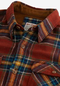 Chemise à boutons en flanelle à carreaux rouge rouille et bleu marine. Présente une texture douce, deux poches avant et un col intérieur beige avec des détails cousus.