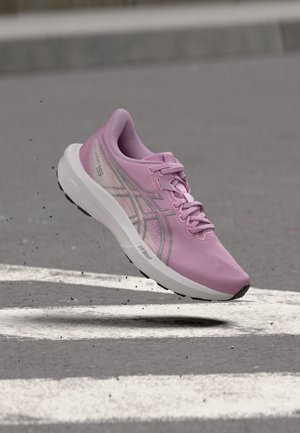 Chaussure de course athlétique rose avec semelle blanche en l'air au-dessus de l'asphalte avec des marquages routiers blancs.