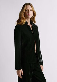 Pikkade blondide juustega naine, kes kannab musta sametist avatud jopet ja sobivaid musti sametist pükse, taustaks hele ühtlane sein.