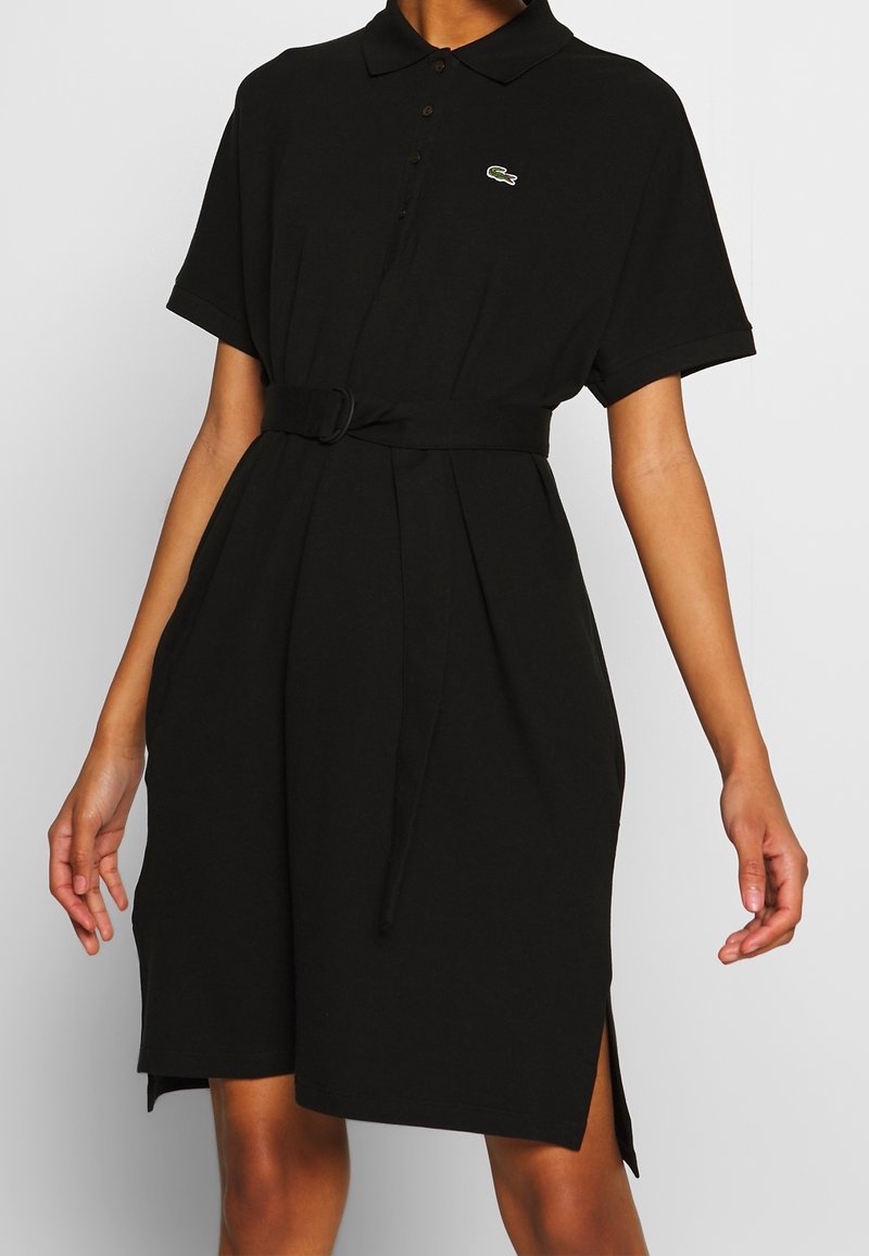 Robe polo noire à manches courtes avec ceinture et fentes latérales, avec un petit logo crocodile vert sur la poitrine.