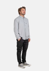 Camisa de botones de manga larga gris con un bolsillo en el pecho, combinada con pantalones negros y zapatillas gris, de pie sobre un fondo neutro.