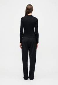 Γυναίκα που στέκεται με την πλάτη γυρισμένη, φορώντας εφαρμοστό μαύρο μακρυμάνικο τοπ και φαρδιά μαύρα παντελόνια, μπροστά σε απλό ανοιχτόχρωμο φόντο.