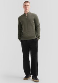 Oliwkowy sweter z zamkiem pod szyję o prążkowanej fakturze, w połączeniu z czarnymi spodniami z korduroy i czarnymi trampkami. Prosty, nowoczesny design.