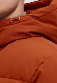 Orange puffer jacket with a hood, featuring a textured fabric and adjustable drawstring. Close-up shows seam details and stitching.