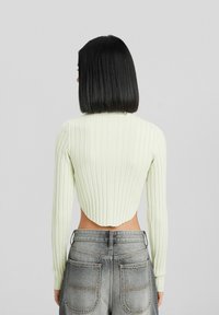 Kvinna med rakt svart hår som bär en ljusgrön ribbad kortärmad tröja och högmidjade grå jeans, vänd bort från kameran mot en enfärgad bakgrund.