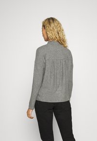 Szary sweter z długimi rękawami, wysokim kołnierzem i marszczonym detalem z tyłu, wykonany z miękkiego, teksturowanego materiału. Połączony z czarnymi spodniami.