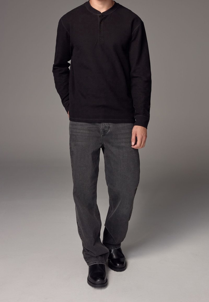 Homme portant une chemise henley noire à manches longues, un jean gris et des bottes noires, debout devant un fond gris uni.