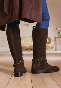 Hnědé semišové kozačky do kolen s texturovaným povrchem, zlatými cvoky a přezkou. Design doplňuje blokový podpatek a přiléhavý tvar.