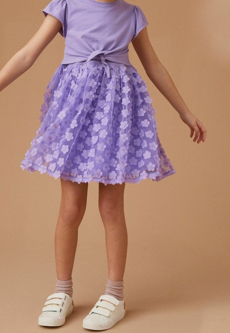 Robe t-shirt violette avec un devant noué et une jupe superposée en tulle lilas ornée d'appliqués en fleurs. Associée à des chaussures de sport blanches.