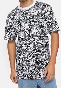 Tričko s krátkým rukávem s černobílým abstraktním vlnkovým vzorem a návrhy usměvavých obličejů; vyrobeno z lehkého materiálu s kulatým výstřihem.