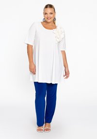Tunique blanche à manches courtes, avec un accent floral en tissu sur l'épaule. Associée à un pantalon bleu ajusté et des sandales blanches.