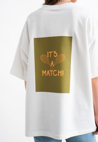 Witte oversized t-shirt met een groene rechthoekige grafische afbeelding met de tekst "HET IS EEN MATCH!" en twee tennisraketten in oranje.