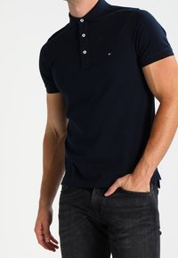 Homme portant un polo à manches courtes bleu marine avec des boutons blancs et un jean noir, la main dans la poche, sur un fond blanc.