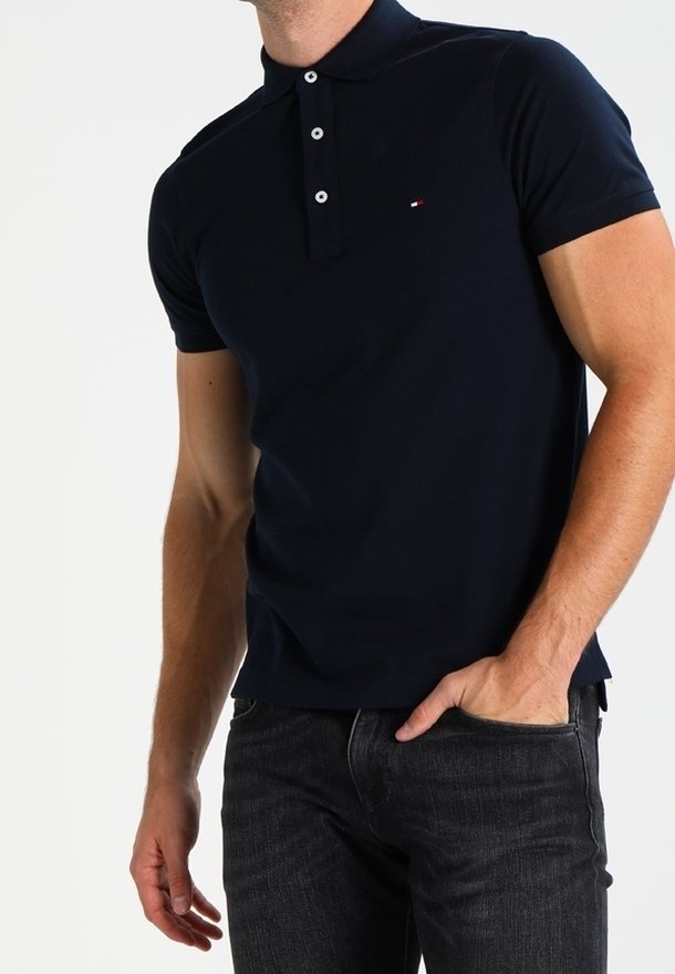 Homme portant un polo à manches courtes bleu marine avec des boutons blancs et un jean noir, la main dans la poche, sur un fond blanc.