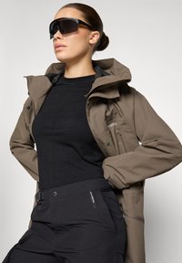 Pruun veekindel jope musta särgi peal, kombineeritud mustade cargo-pükstega. Suured mustad päikeseprillid ja sile, moodne disain.