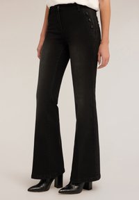 Schwarze, weit ausgestellt Jeans mit hoher Taille, mit einem geknöpften Seitendetail und einem glatten, leicht strukturierten Stoff. Getragen mit Absatzstiefeln im krokodilartigen Muster.