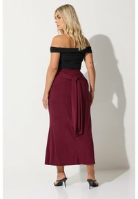 Asymmetrisk svart topp med axlarna bar, tillsammans med en figurnära bordeaux långkjol som har en knytning vid midjan och en slät, något glänsande yta.