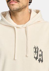 Baardman die een crèmekleurige hoodie draagt met een zwart gotisch "VA" logo op de linkerborst en zichtbare koorden.