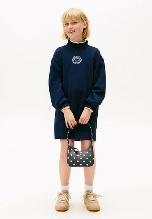 Robe sweatshirt marine avec col volant et motif de cœur brodé. Sac à main à pois noirs. Baskets beiges avec des lacets clairs.