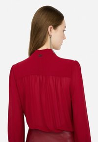 Blusa rossa a maniche lunghe con una texture morbida, caratterizzata da uno schienale plissettato e un accento di bottone al collo.