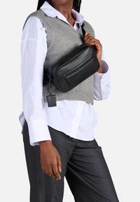 Borsa a tracolla in pelle nera con logo impresso, chiusura a zip e dettagli testurizzati. Indossata in stile crossbody sopra un gilet grigio in maglia su una camicia bianca.