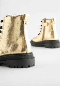 Goldfarbene, metallische Knöchelstiefel mit strukturiertem Finish, schwarzen Schnürsenkeln, einem seitlichen Reißverschluss und einer klobigen Gummisohle mit ausgeprägtem Profil.