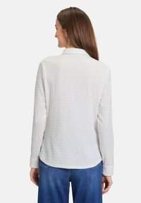 White textured long-sleeve shirt with a collar, featuring a relaxed fit and curved hem, paired with blue jeans.