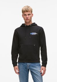 Sudadera negra de mezcla de algodón, con un bolsillo delantero, capucha con cordón y logo azul en el pecho. Combinada con jeans de mezclilla desgastados.