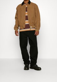 Chaqueta marrón con cremallera y cuello negro, combinada con una camiseta de rayas en color burdeos y amarillo, acompañada de pantalones negros y zapatos negros.