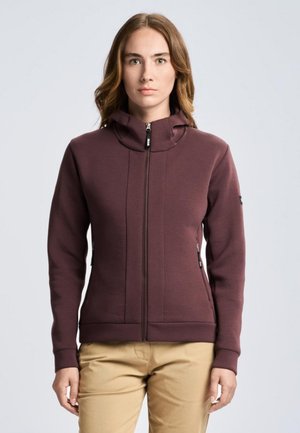 Una mujer luce una chaqueta color ciruela con cremallera y cuello alto, y unos pantalones beige, sobre un fondo liso y claro.