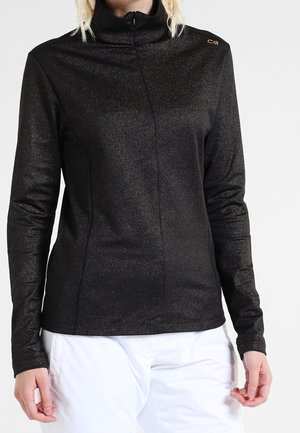 Frau trägt ein langärmliges, schwarzes, schimmerndes Zip-up-Oberteil mit hohem Kragen und weiße Hose, steht vor einem schlichten Hintergrund.