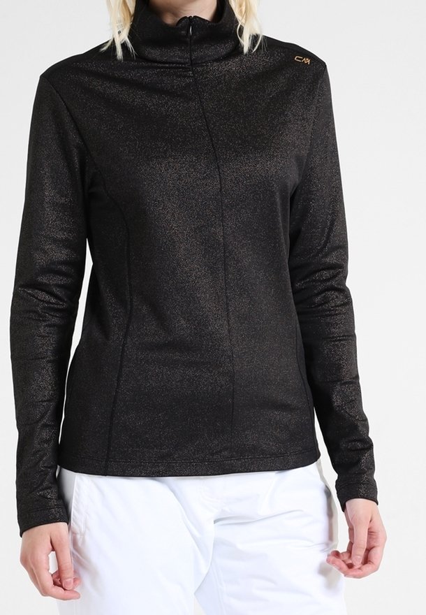 Femme portant un haut zippé noir scintillant à manches longues avec un col montant et un pantalon blanc, debout devant un fond uni.