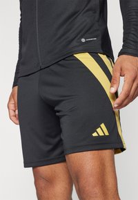 Schwarze Sportshorts mit gelben diagonalen Streifen, aus glattem Material hergestellt. Verfügt über ein Adidas-Logo auf der unteren linken Seite.