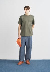 Χακί βαμβακερό μπλουζάκι με μικρό λογότυπο, φαρδιά μπλε τζιν παντελόνια, πορτοκαλί καπέλο κρατημένο στο χέρι και πορτοκαλί παπούτσια με κορδόνια.