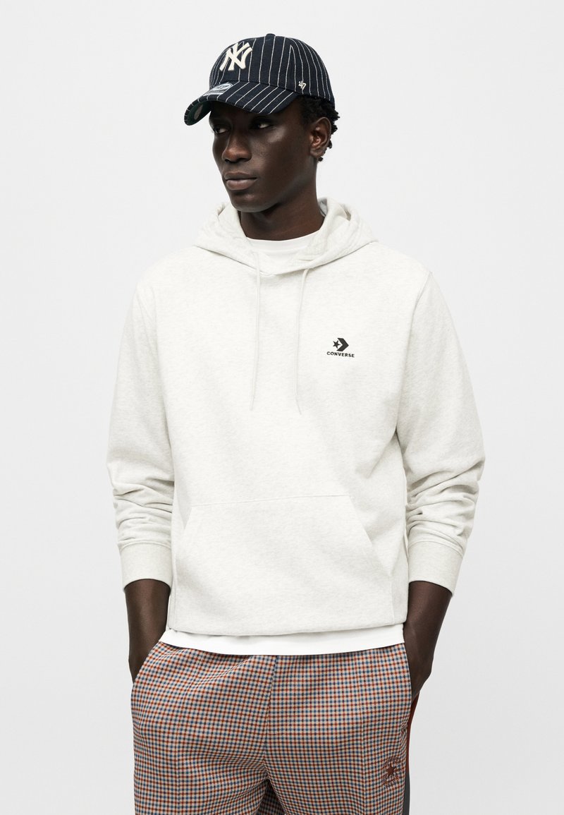 Sweat à capuche gris clair avec une poche avant et un logo noir ; assorti à un pantalon à carreaux orange et bleu ; casquette rayée noire et blanche.