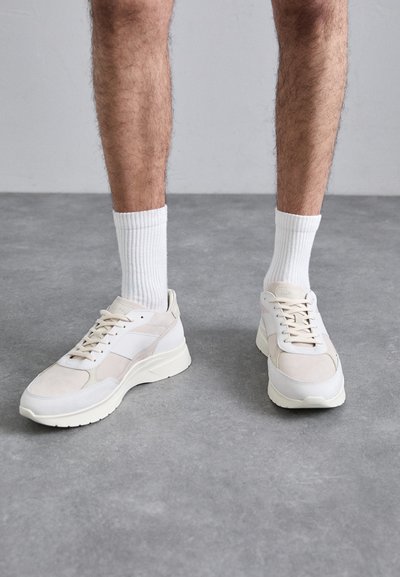 Adidași bej deschis cu un amestec de materiale netede și texturate, accente albe, închidere cu șireturi și o talpă în contrast.