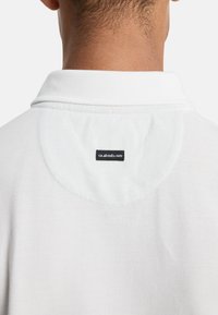 Λευκό πόλο μπλουζάκι με υφή υφάσματος, κλασικό γιακά και στρογγυλό γιόκ στο πίσω μέρος με μια μικρή μαύρη ετικέτα με λογότυπο.