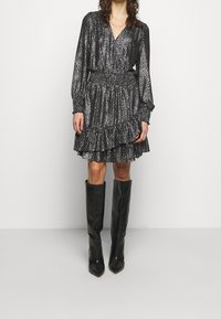 Robe noire et argentée à pois avec manches longues, col en V et ourlet à volants superposés, associée à des bottes noires pointues montant jusqu'aux genoux.