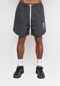 Pantaloni corti neri con vita elasticizzata e cordino beige, con logo sulla gamba sinistra. Il materiale appare leggero e strutturato.